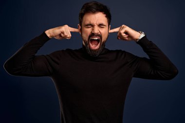 Çaresiz sakallı adamın fotoğrafı ellerini kafasından ayırmıyor, yüksek sesle bağırıyor, kulaklarını parmaklarıyla kapatıyor, ağzını açıyor, gözlerini kapatıyor, depresif hissediyor, borçları ve finansal sorunları var, süveter giyiyor..