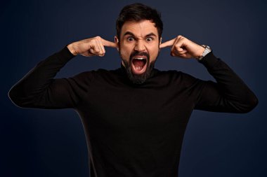 Çaresiz sakallı adamın fotoğrafı ellerini kafasından ayırmıyor, yüksek sesle bağırıyor, kulaklarını parmaklarıyla kapatıyor, ağzını açıyor, gözlerini kapatıyor, depresif hissediyor, borçları ve finansal sorunları var, süveter giyiyor..