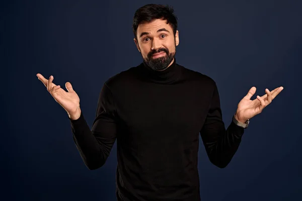 Carefree, indecisive young handsome businessman in black high neck sweater, shrugging raise hands sideways, tilt head and smiling as apoligizing, cant help, not know answer, standing Blue background.