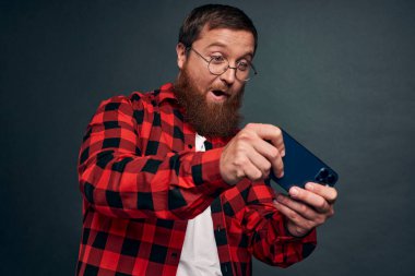 Ekose gömlekli heyecanlı sakallı adam gri arka planda akıllı telefonla oynuyor..