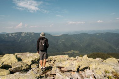 Sırt çantalı genç bir adam bir dağın tepesinde duruyor.
