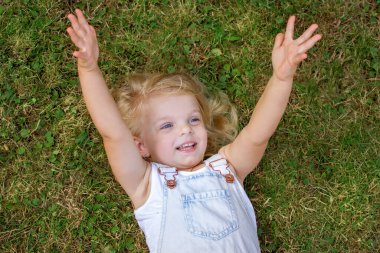 Yeşil çimlerin üzerinde uzanan mutlu sarışın kız ellerini kaldırıyor ve samimi bir şekilde gülümsüyor. Mutlu çocukluk.