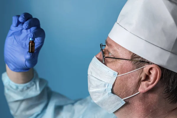 An elderly doctor in medical clothing: a cap, gloves,dressing gown and mask, glasses. Examines a bottle of medicine or vaccine