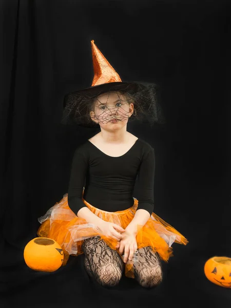 A girl in a witch costume - a hat with a spider web and an orange skirt-sits wearily on a black background. Next to it is a bucket-pumpkin for collecting sweets. Waiting for the holiday