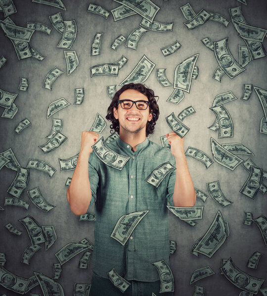 Contented young man keeps fists tight raised up as money are falling like a dollar rain. Surprised grateful guy celebrate positive expression. Lottery win, unexpected success, wealth and luck concept.