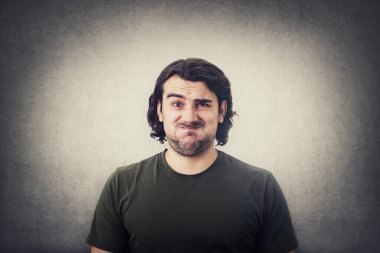 Şaşkın ve rahatsız olmuş genç bir adamın portresi, uzun kıvırcık saçlı, kameraya kafası karışmış ve hayal kırıklığına uğramış görünüyor, yanaklarını üflüyor, kaşlarını çatıyor ve yüzünü ekşitiyor. Gri duvarda izole edilmiş mutsuz bir adam.