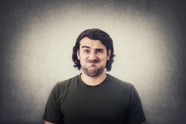 Portrait of bewildered and annoyed young man, long curly hair, looking baffled and frustrated to camera, blowing his cheeks, frowning and making perplexed grimace. Displeased guy isolated on grey wall clipart