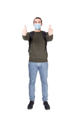 Koronavirüsten korunmak için yüz maskesi takan ihtiyatlı bir gencin portresi beyaz arka planda iki başparmağın da izole olduğunu gösteriyor. Sıradan erkek güvenliği, COVID-19 önleme tedbirleri.