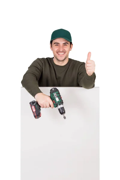 ドライバーや電動ドリルを手にした積極的な改修作業員は 白いパネルの後ろに立って 親指を上げてジェスチャーを示します 内装仕上げ板やDiy家具を組み立てる男 — ストック写真