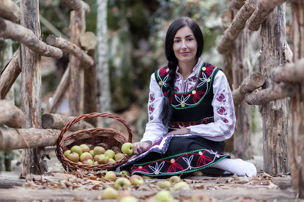 Молодая девушка в болгарском костюме сидит на земле с корзиной с яблоками — стоковое фото