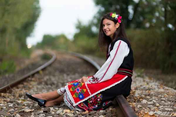 Красивая молодая девушка в болгарском костюме сидит на железнодорожной линии — стоковое фото