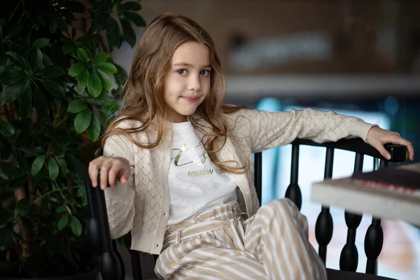 Cute white teenage girl with long wavy hair sits near the window at a table indoors with wooden walls and thinks about something, smiles — Stock Photo, Image