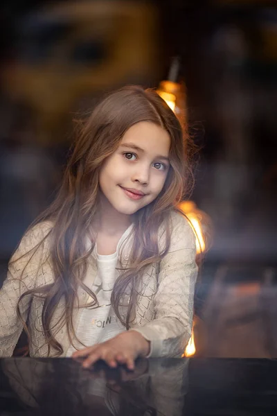 Cute white teenage girl with long wavy hair sits near the window at a table indoors with wooden walls and thinks about something, smiles — Stock Photo, Image