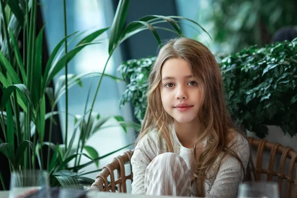 Schattig wit tienermeisje met lang golvend haar zit bij het raam aan een tafel binnen met houten muren en denkt aan iets, glimlacht — Stockfoto