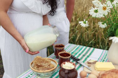  Beyaz elbiseli hamile bir kızın elleri bardağa taze süt döker. Tatlım, yumurta, tereyağı. Buğday tarlası.