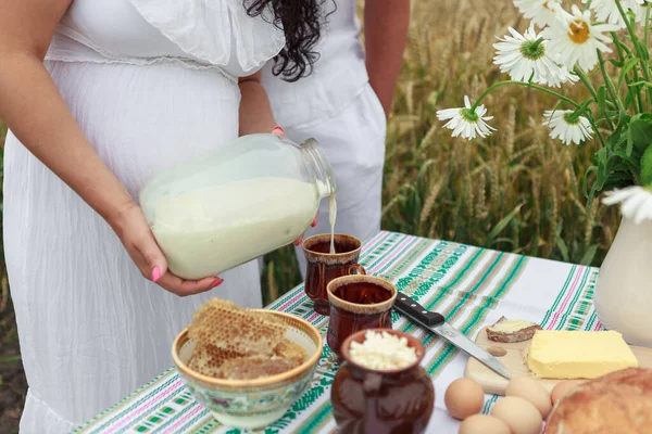 Hände eines schwangeren Mädchens im weißen Kleid gießen frische Milch aus einer Dose in einen Becher. Honig, Eier, Butter auf dem Tisch. Weizenfeld. Stockbild