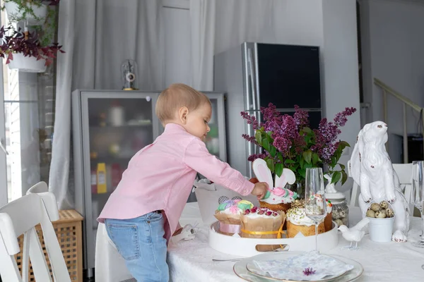 Kleine schattige witte jongen staat aan de paastafel en het eten van paastaart, lila, paashaas, Pasen decoraties, geschilderde paaseieren. — Stockfoto