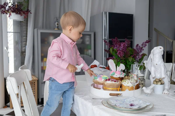 Piccolo ragazzo bianco carino è in piedi al tavolo di Pasqua e mangiare torte pasquali, lilla, coniglietto di Pasqua, decorazioni pasquali, uova di Pasqua dipinte. — Foto Stock