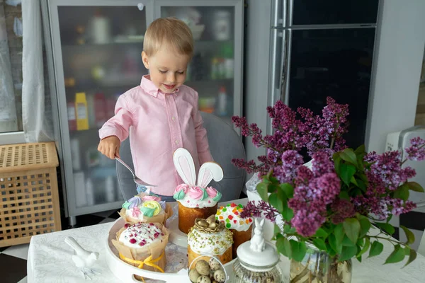 Piccolo ragazzo bianco carino è in piedi al tavolo di Pasqua e mangiare torte pasquali, lilla, coniglietto di Pasqua, decorazioni pasquali, uova di Pasqua dipinte. — Foto Stock