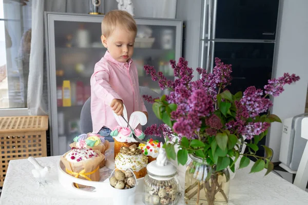 Маленький милый белый мальчик стоит за пасхальным столом и ест пасхальные торты, сирень, пасхальный кролик, пасхальные украшения, окрашенные пасхальные яйца. — стоковое фото