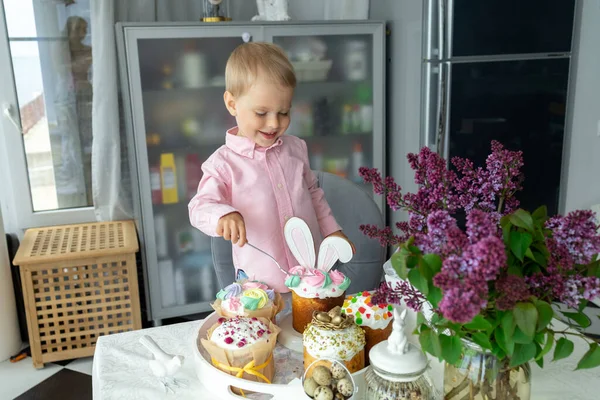 Piccolo ragazzo bianco carino è in piedi al tavolo di Pasqua e mangiare torte pasquali, lilla, coniglietto di Pasqua, decorazioni pasquali, uova di Pasqua dipinte. — Foto Stock