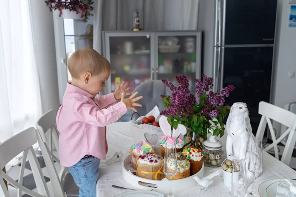 Kleine schattige witte jongen staat aan de paastafel en het eten van paastaart, lila, paashaas, Pasen decoraties, geschilderde paaseieren. — Stockfoto