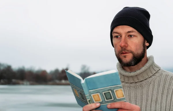 Portrait young man in sweater with cap reading book in winter Czech landscape