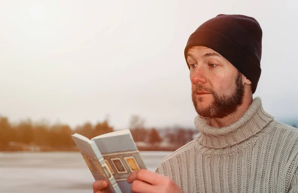 Portrait young man in sweater with cap reading book in winter Czech landscape from side