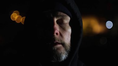 Kapüşonlu sakallı adamın gece kapanış portresi. Arka planda ışıklar var. Meditasyon, rahatlama konsepti