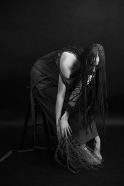 Siyah-beyaz Stüdyo fotoğrafı, uzun siyah elbiseli, afro örgülü, bitkilerle poz veren genç bir kız.