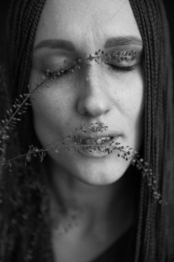 Siyah-beyaz Stüdyo fotoğrafı, uzun siyah elbiseli, afro örgülü, bitkilerle poz veren genç bir kız.