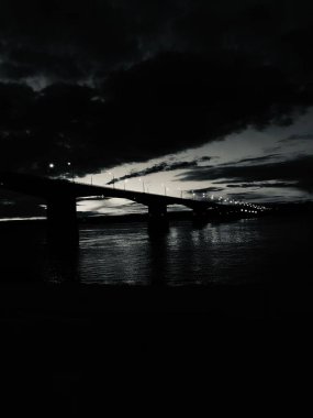 Nehir üzerindeki bir köprünün siyah beyaz fotoğrafı, karanlık şehir manzarası.