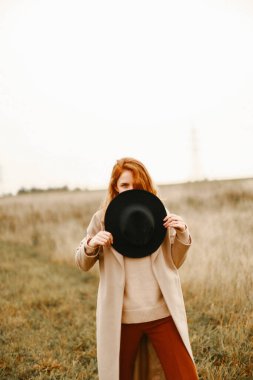 Sonbahar portresi, siyah şapkalı kızıl saçlı bir kız ve doğada yürüyen kahverengi bir ceket. Doğal yerlerdeki güzel portre, bulanık arkaplan ve bulanık odak