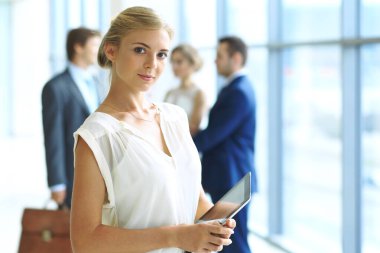 Dokunmatik bilgisayarlı sarışın kadın kameraya bakıyor ve gülümsüyor iş adamları arka planda el sıkışırken.