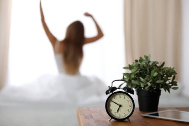 Alarm clock standing on bedside table has already rung early morning to wake up woman is stretching in bed in background. Early awakening, not getting enough sleep, oversleep concept
