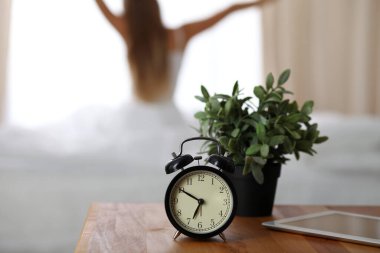 Alarm clock standing on bedside table has already rung early morning to wake up woman in bed sitting in background. Early awakening, not getting enough sleep, oversleep, time line concept