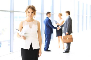 Dokunmatik bilgisayarlı sarışın kadın kameraya bakıyor ve gülümsüyor iş adamları arka planda el sıkışırken.