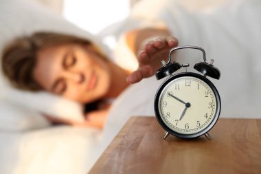 Uykucu genç kadın alarmın çalmasına elini uzatıyor ve kapatıyor. Erken kalkmak, uykusuzluk, iş kavramı..
