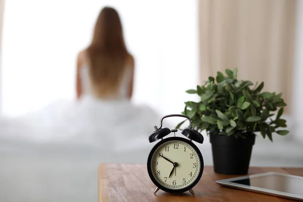 Alarm clock standing on bedside table has already rung early morning to wake up woman is stretching in bed in background. Early awakening, not getting enough sleep, oversleep concept — 스톡 사진