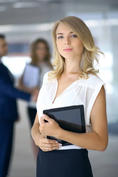 Ξανθιά γυναίκα με touchpad υπολογιστή κοιτάζοντας κάμερα και χαμογελώντας, ενώ οι επιχειρηματίες χειραψία πάνω από το παρασκήνιο — Φωτογραφία Αρχείου