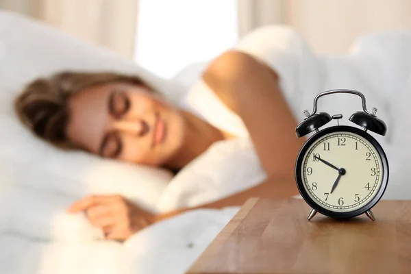 Beautiful young woman sleeping and smiling while lying in bed comfortably and blissfully on the background of alarm clock is going to ring. Sunbeam dawn on her face. — Stock Photo, Image