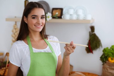 Çekici, genç, siyah saçlı bir kadın mutfak sobasının yanında durup gülümserken tahta kaşıkla sıcak yemek tadıyor. Yemek pişirme ve ev idaresi kavramları.