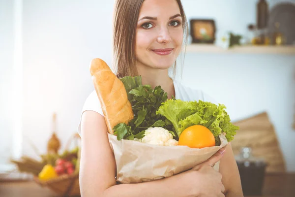 An attractive young woman holding the paper bag full of vegetables while standing and smiling in sunny kitchen. Cooking concept.