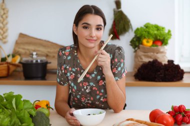 Çekici, genç, koyu saçlı bir kadın mutfakta oturup gülümserken lezzetli bir salata karışımı için yeni bir tarif tadıyor. Yemek pişirme ve ev idaresi kavramları.
