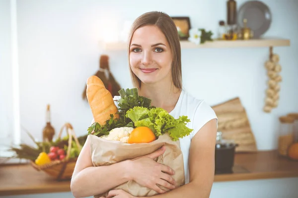 An attractive young woman holding the paper bag full of vegetables while standing and smiling in sunny kitchen. Cooking concept.