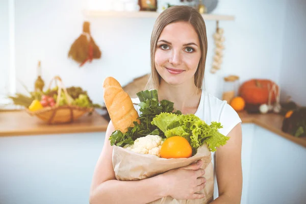 An attractive young woman holding the paper bag full of vegetables while standing and smiling in sunny kitchen. Cooking concept.