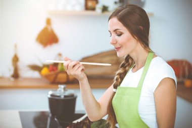 Çekici genç bir kadın, güneşli mutfakta ayakta durup gülümserken yeni keto tarifiyle çorba hazırlıyor. Yemek pişirme ve ev idaresi kavramları.