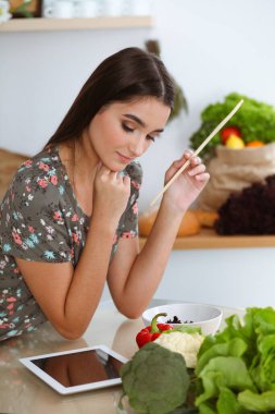 Çekici, genç, koyu saçlı bir kadın mutfakta masada otururken lezzetli bir salata karışımı için yeni bir tarif tadıyor. Tablet pc en iyi yemek kitabıdır.