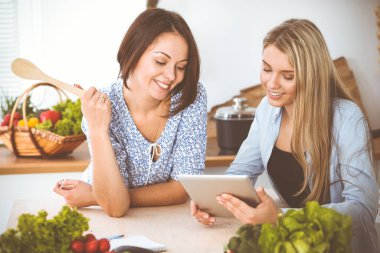 Mutfakta masada otururken iki kadın arkadaş lezzetli bir yemek tarifini seçiyor. Tablet pc en iyi yemek kitabıdır.