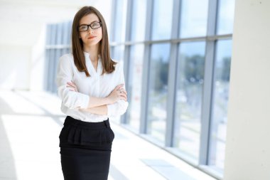 Kollarını kavuşturup modern ofiste duran ve büyüleyici bir şekilde gülümseyen güzel bir kadın uzman. Tasarım, veri analizi, plan stratejisi üzerinde çalışıyorum. İş adamları konsepti.